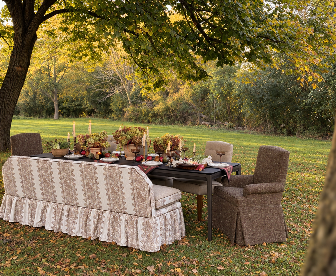 A long dining table sits in an open field, decorated with dried florals. The seating arrangement is complete with a custom banquette, boucle captain's chairs, and four traditional dining chairs.